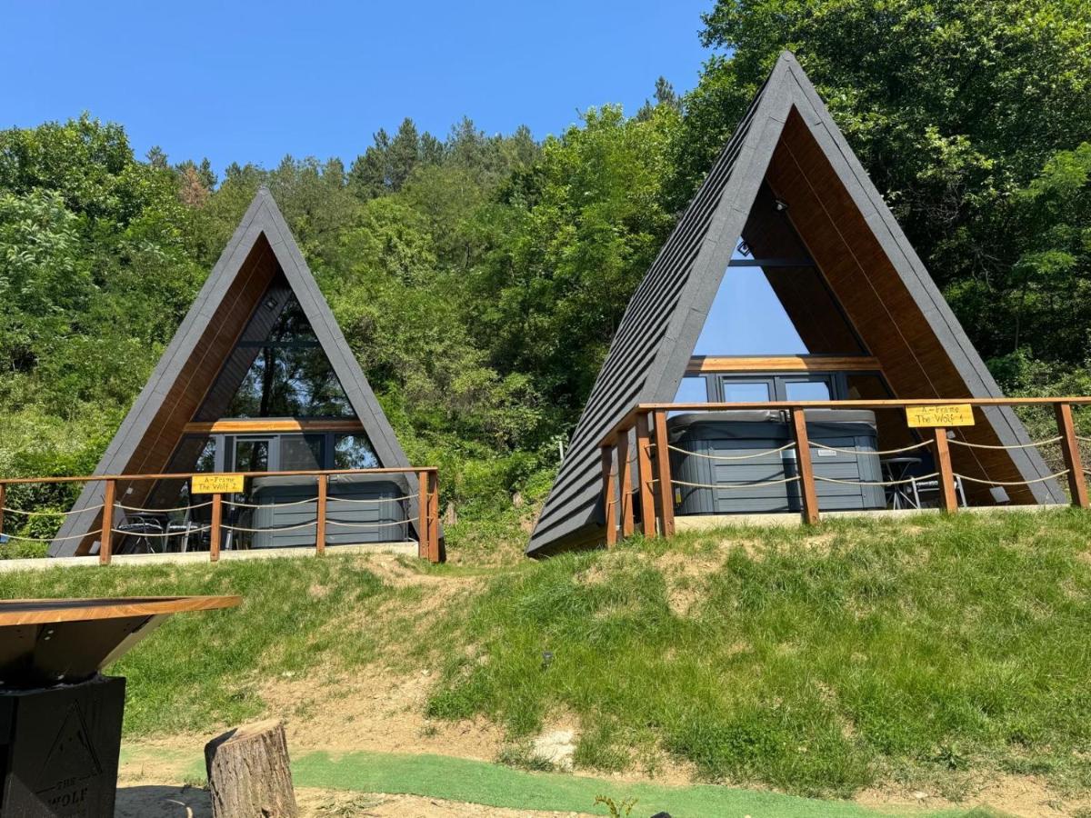 Cele două cabane A-frame așezate una lângă alta pe pajiște, fiecare cu terasă și ciubăr propriu